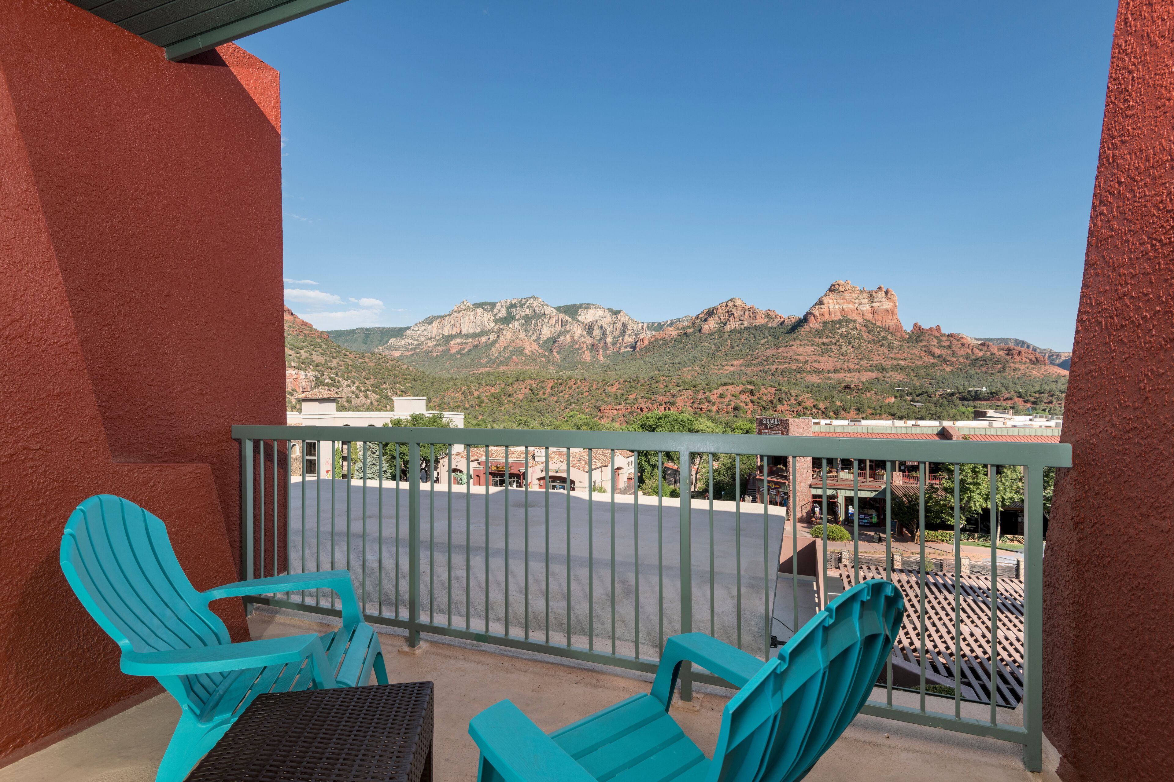 A balcony with turquoise chairs and a table, overlooking a parking area and mountains.