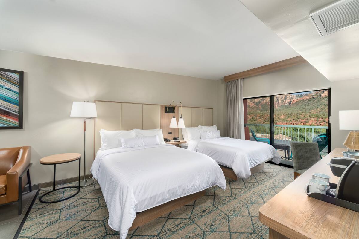 A hotel room with two beds, a desk, a chair, a lamp, and a view of the outdoors.