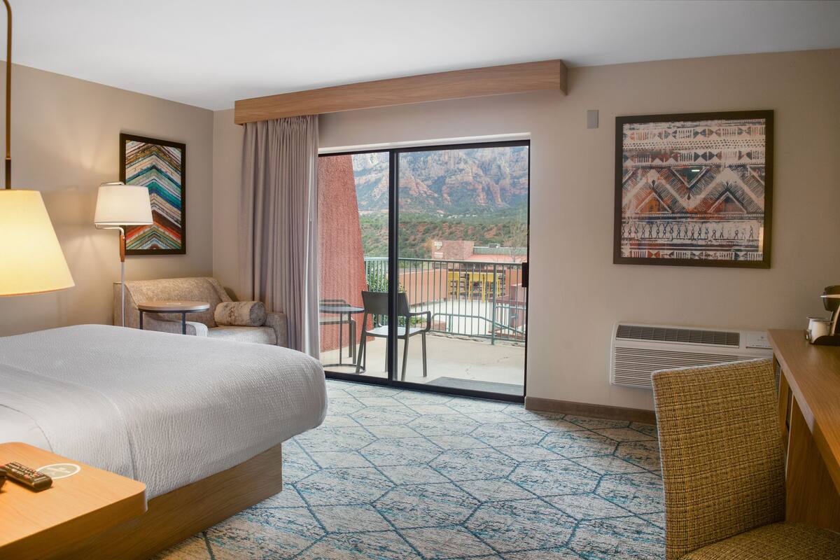 A hotel room with a bed, a desk, a chair, a table, a lamp, a window, and a view of mountains.