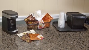 A coffee station with a coffee maker, a basket of coffee pods, and disposable cups on a granite countertop. - The Bluebird Motel (Machias)