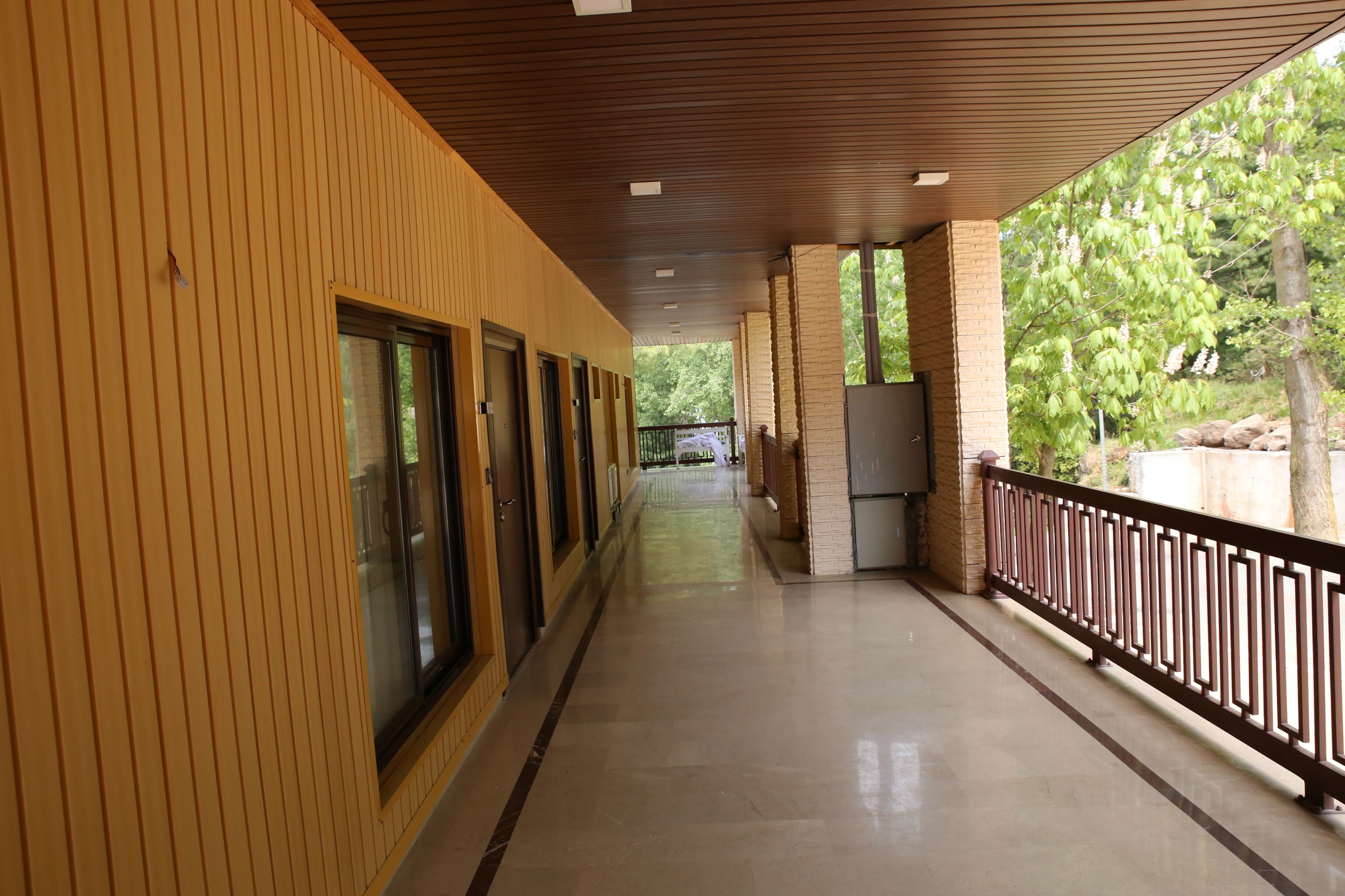 Um corredor coberto com parede de madeira, chão de azulejos e guarda-corpo de metal, com vista para uma área ajardinada com árvores e pedras.