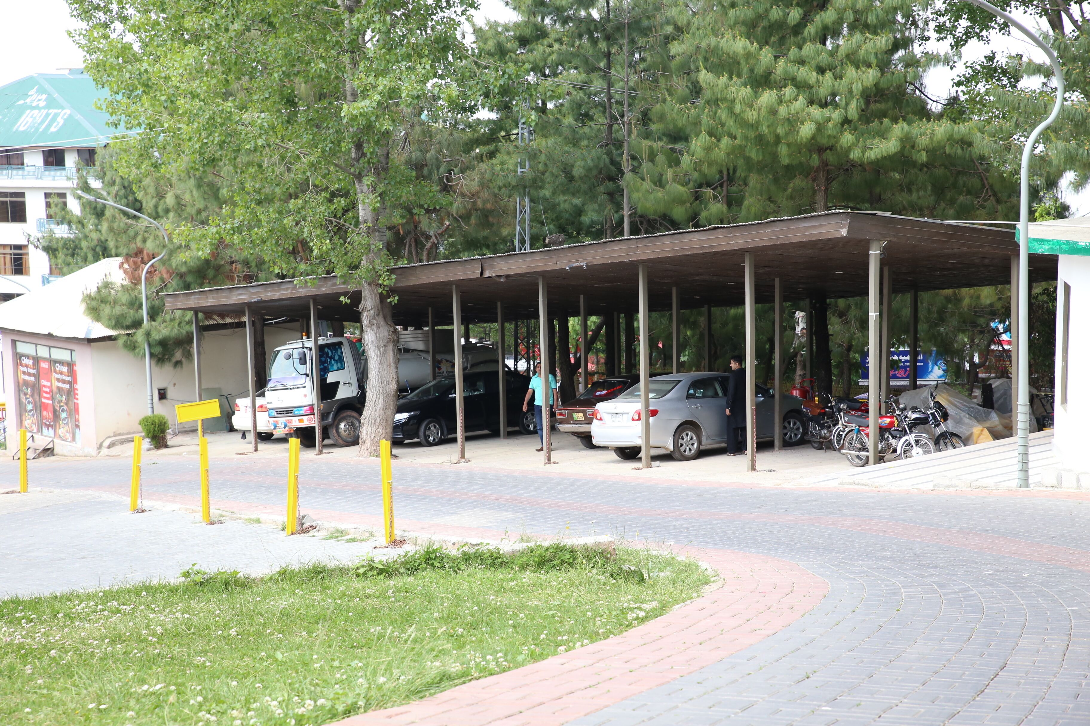 A covered parking area with cars and a truck, surrounded by trees and a building with a green roof.
