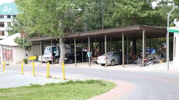 A covered parking area with cars and a truck, surrounded by trees and a building with a green roof.