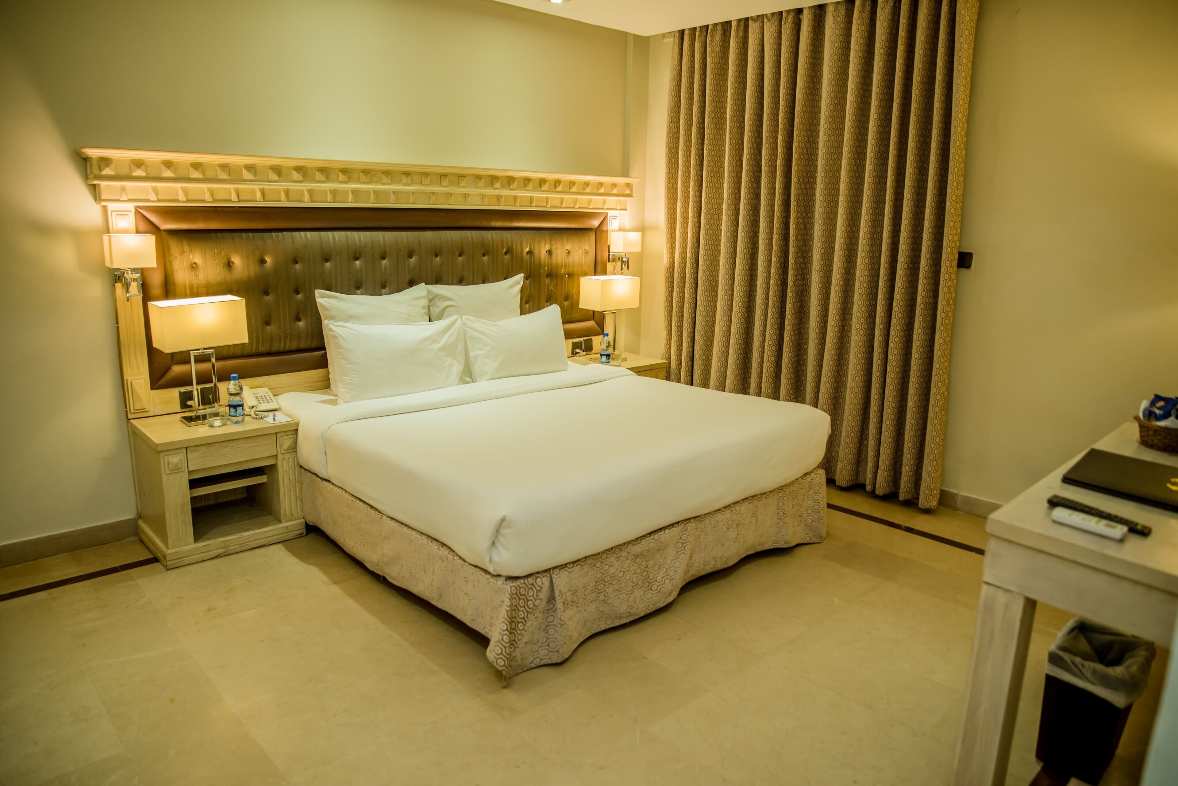 Habitación de hotel con una cama grande, dos mesitas de noche con lámparas, un cabecero y una cortina.