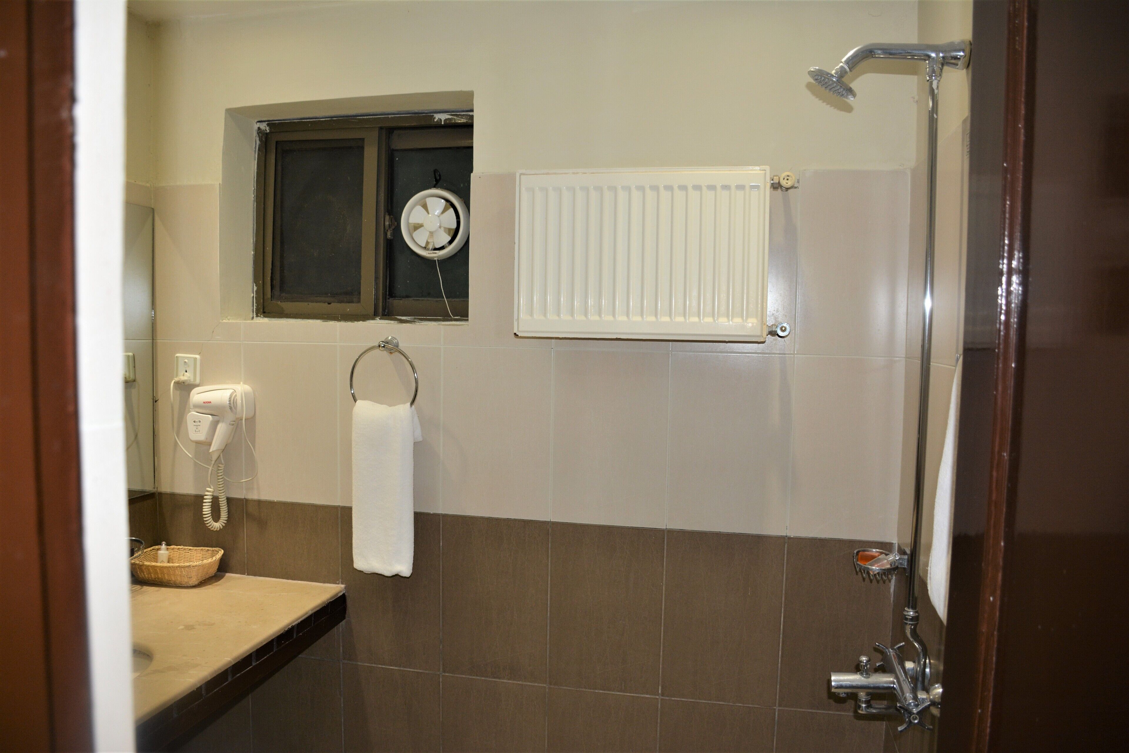 A bathroom with a shower, a hand dryer, a towel rack, and a window with a fan.