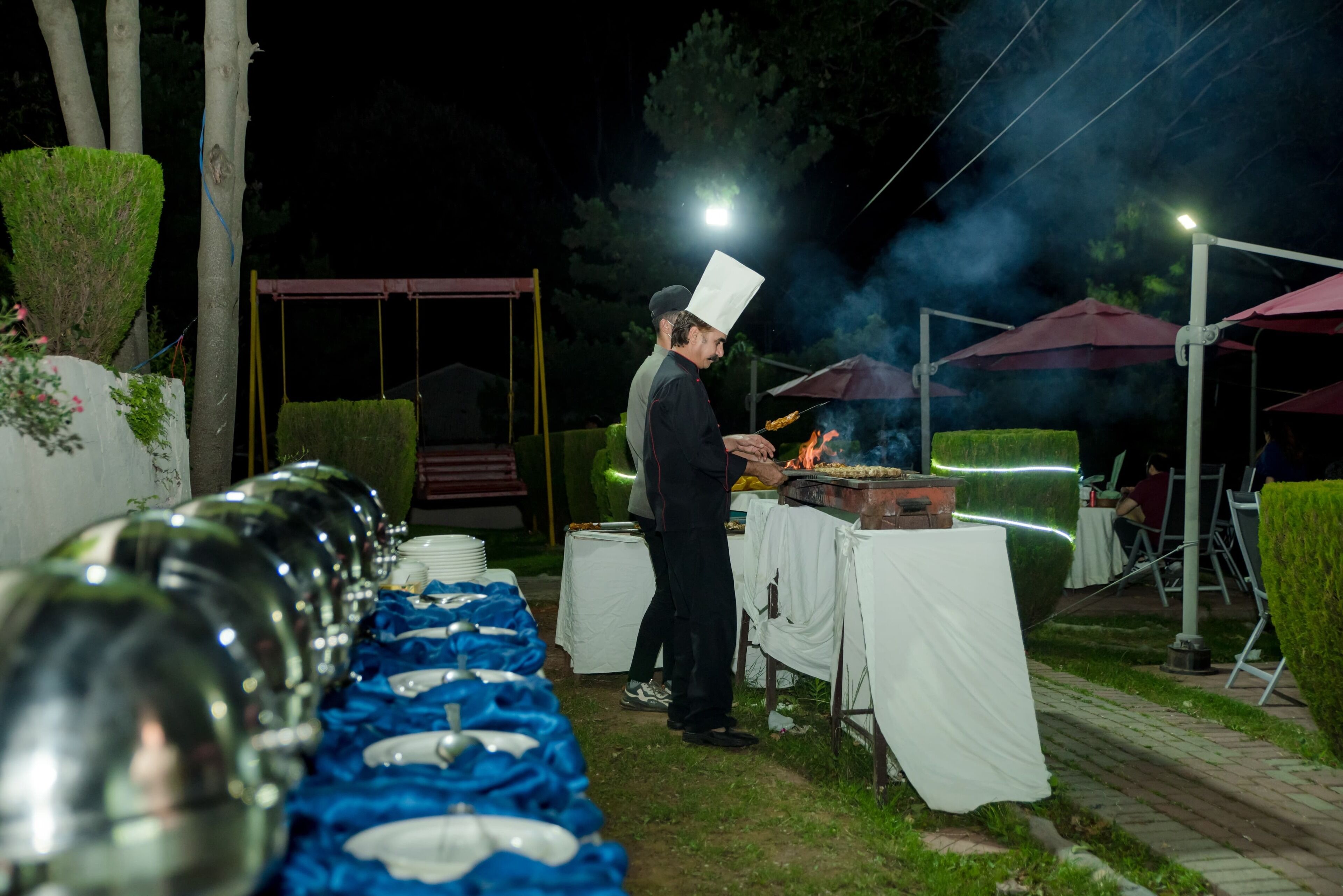 A chef preparing food outdoors at night with a grill, tables with white tablecloths, and a row of glassware.