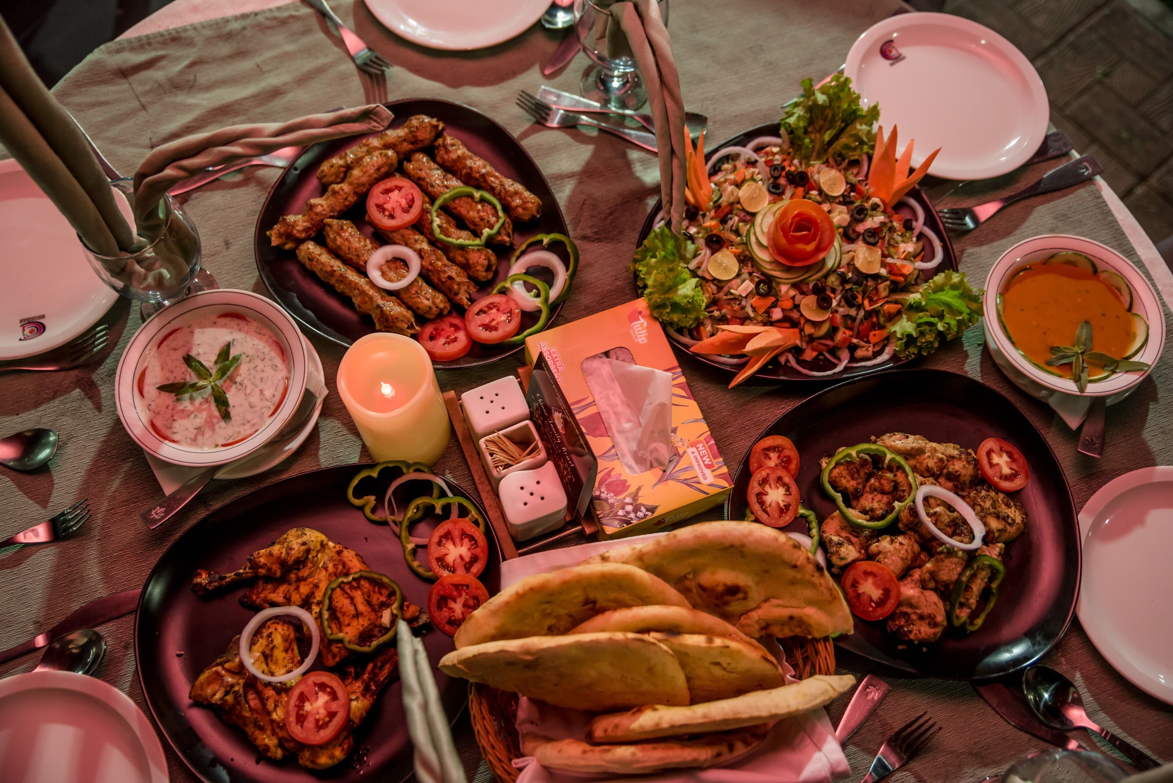 A table set with various dishes including kebabs, salads, and bread, with a candle and a menu.