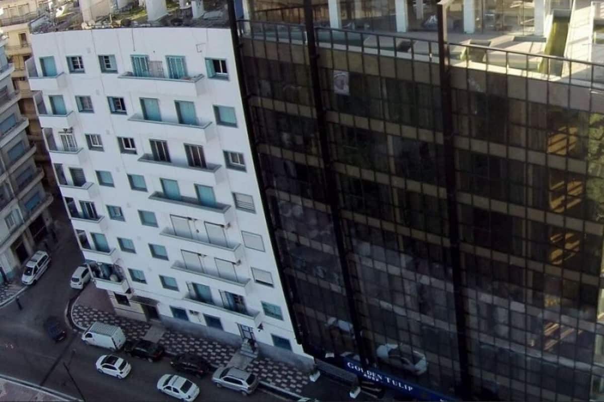 Un immeuble de grande hauteur avec une façade en verre, un bâtiment blanc adjacent doté de balcons, et des voitures garées dans la rue.