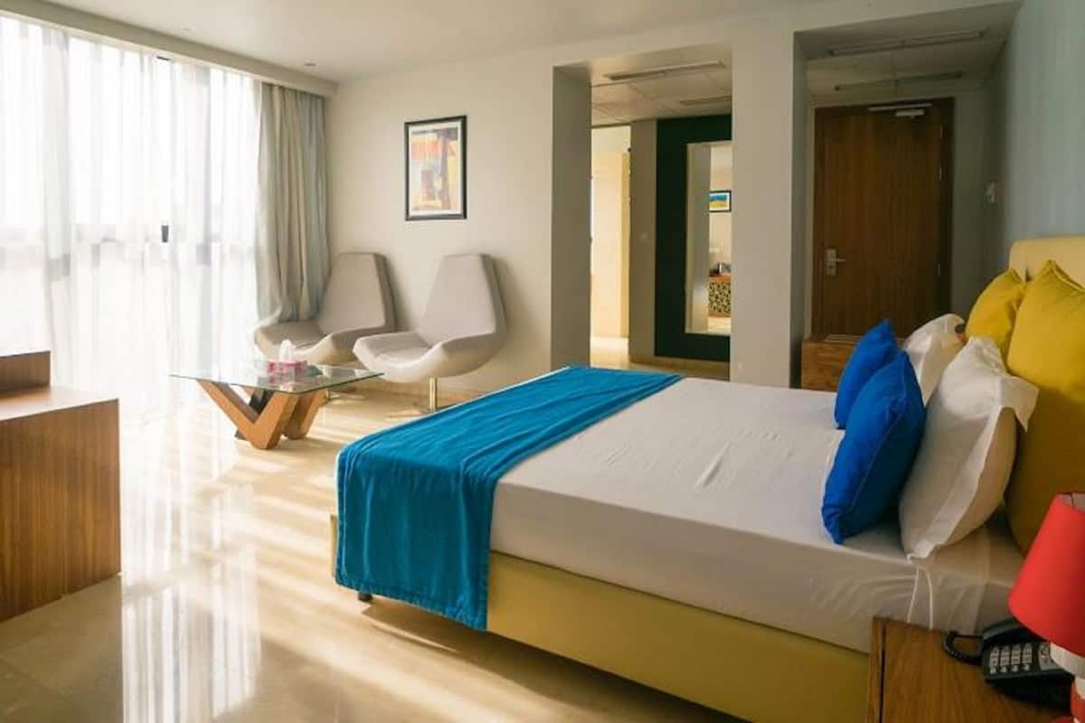 Une chambre d’hôtel comprenant un lit, un bureau, deux chaises, une table en verre et une porte donnant sur une autre pièce.