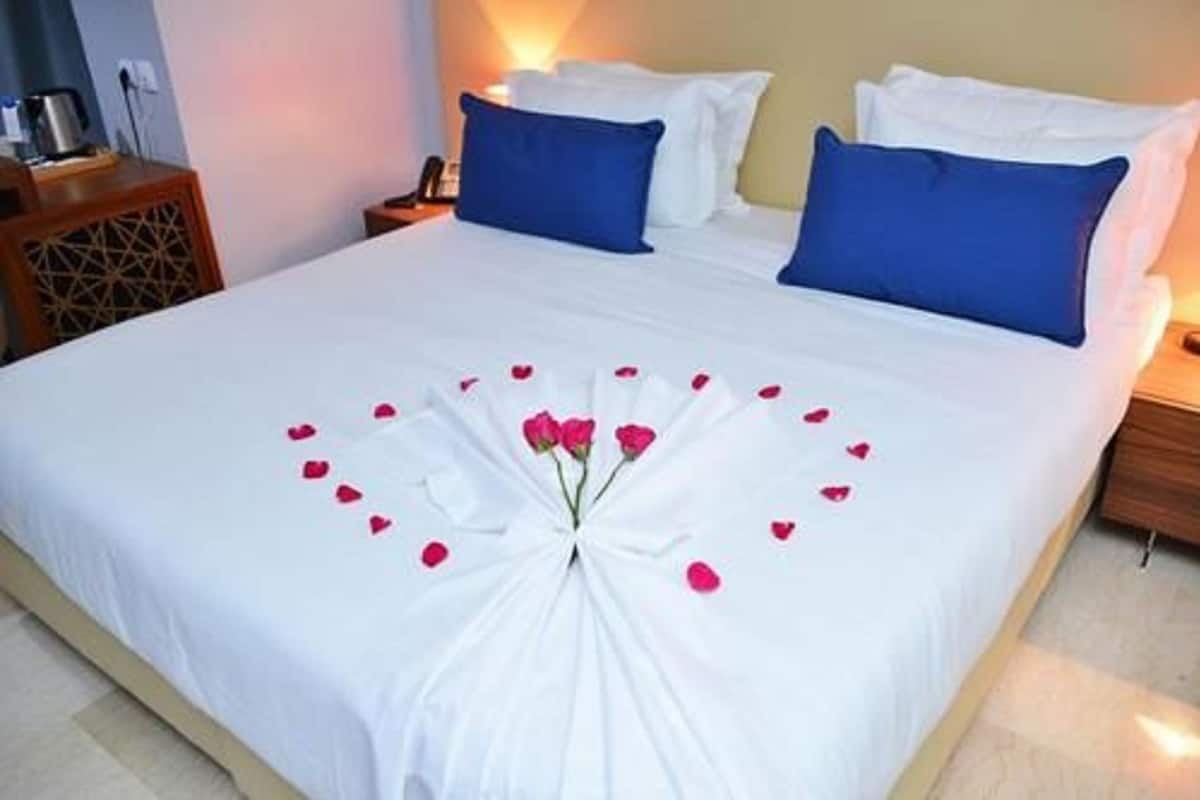 Un lit bien fait, avec des draps blancs, des oreillers bleus et une composition décorative de pétales de rose et un bouquet de roses au centre.