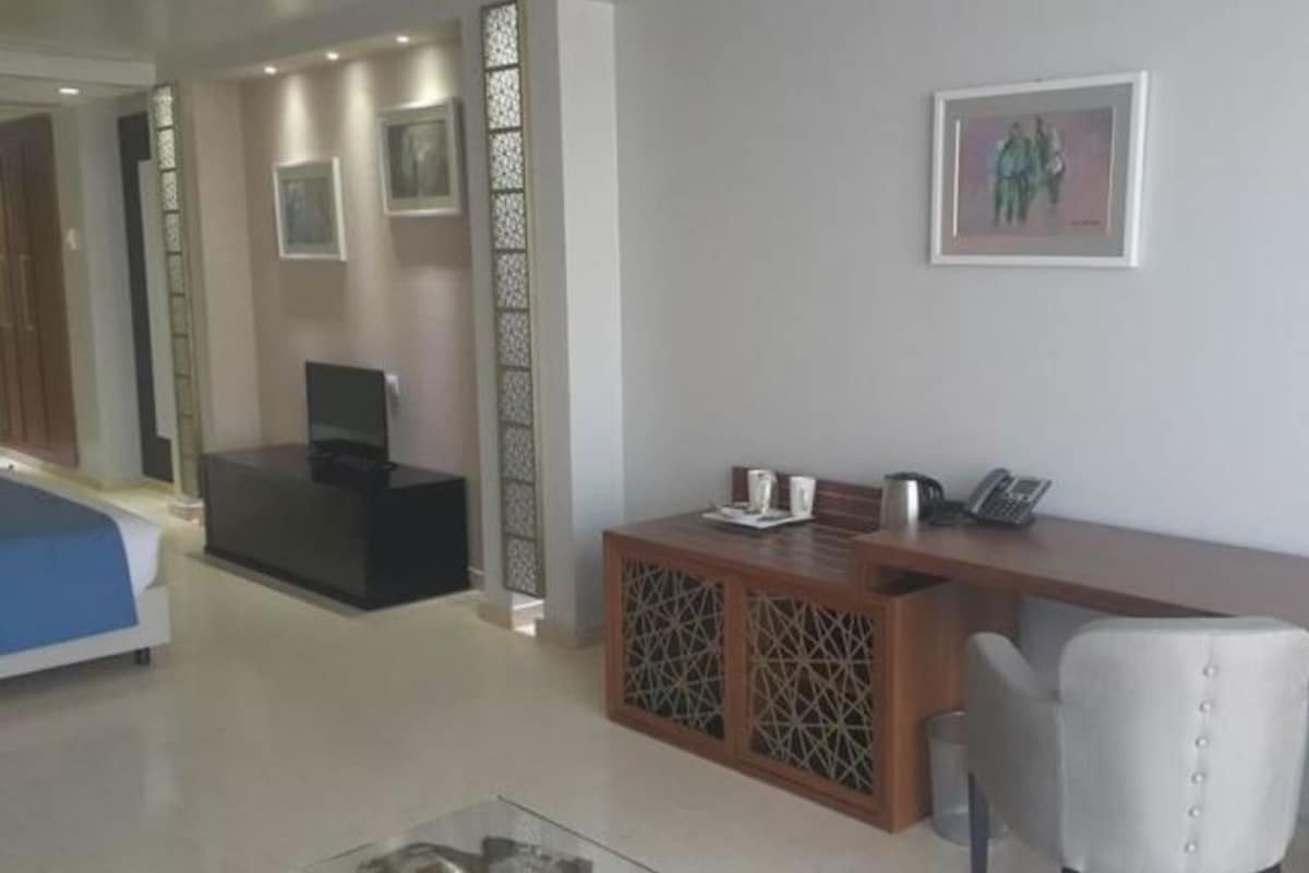 Zona de recepción de hotel con mostrador, un espacio de descanso con una silla y una cama al fondo.