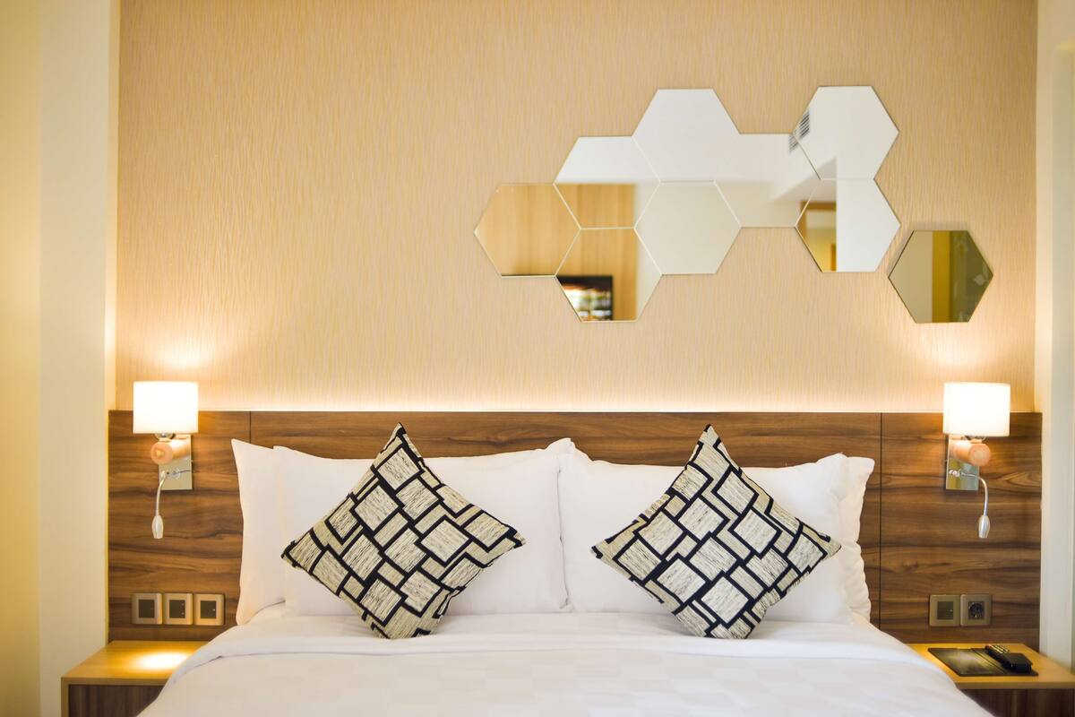 A hotel room with a wooden headboard, two bedside lamps, a bed with white bedding, and a geometric wall mirror.