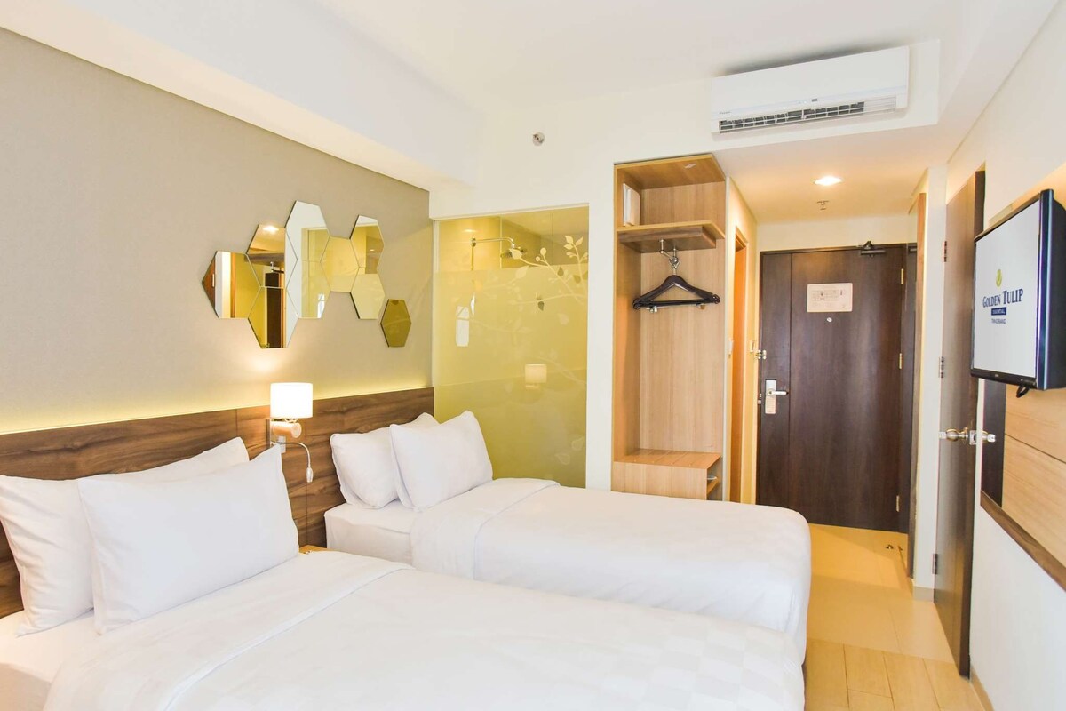 A hotel room with two beds, a mirror, a wall-mounted air conditioner, and a television.