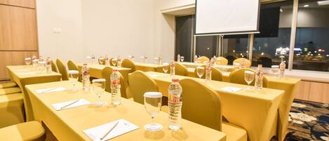 Un salón de conferencias con mesas dispuestas para reuniones, equipado con botellas de agua y vasos, y una pantalla de proyector.