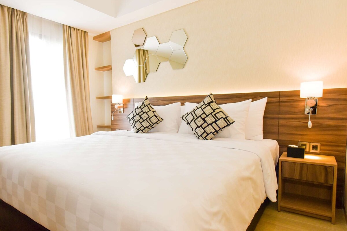 A hotel room with a large bed, two bedside tables, a wall-mounted mirror, and a window with curtains.