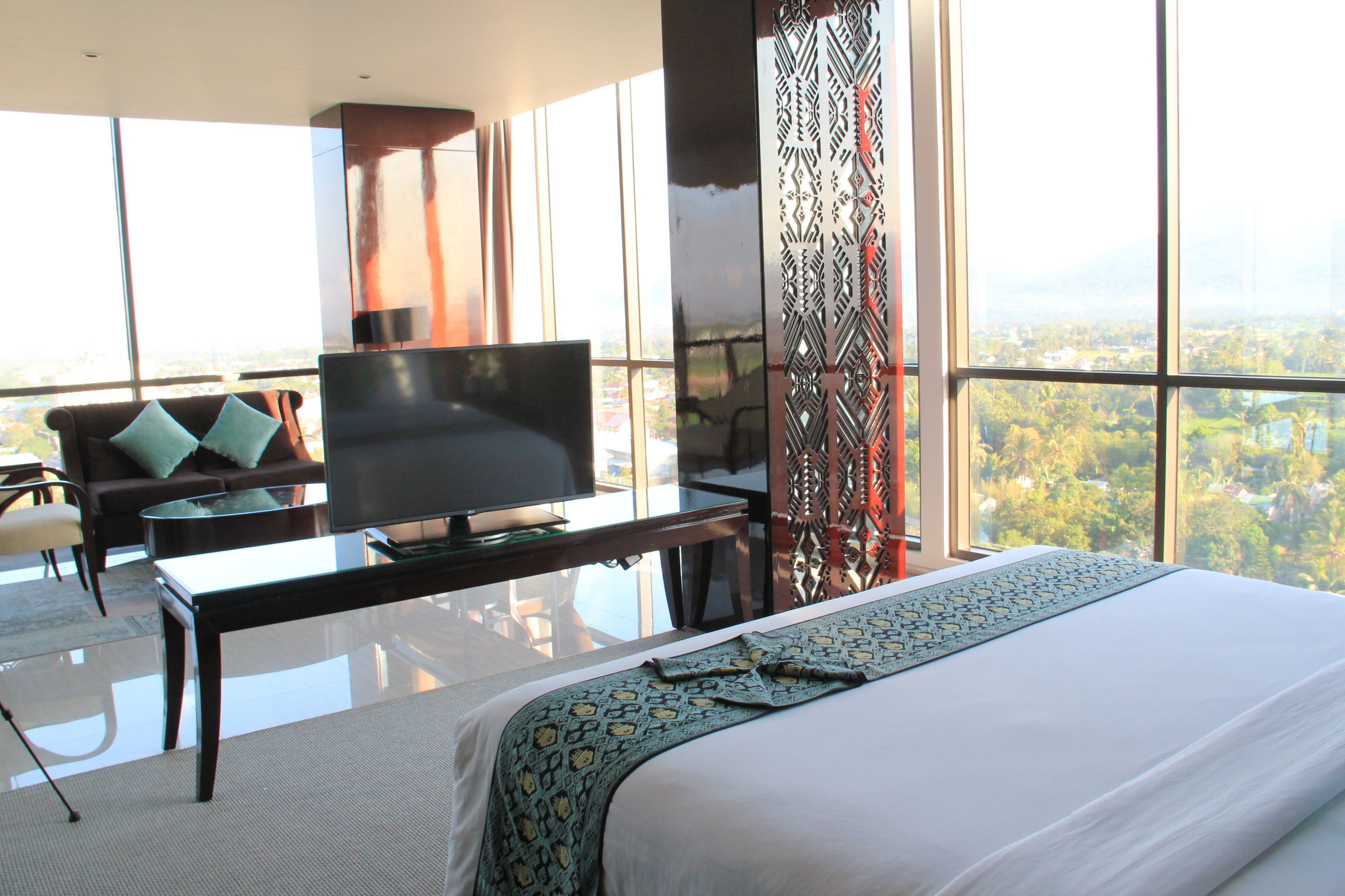 A hotel room with a large bed, a flat-screen TV, a sofa, and a view of a cityscape through large windows.