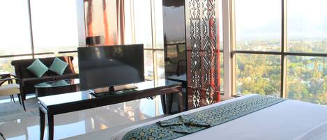 A hotel room with a large bed, a flat-screen TV, a sofa, and a view of a cityscape through large windows.