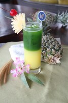 Um copo alto com uma bebida em camadas, decorado com cereja, fatia de abacaxi e uma flor, sobre uma mesa com um abacaxi e flores.