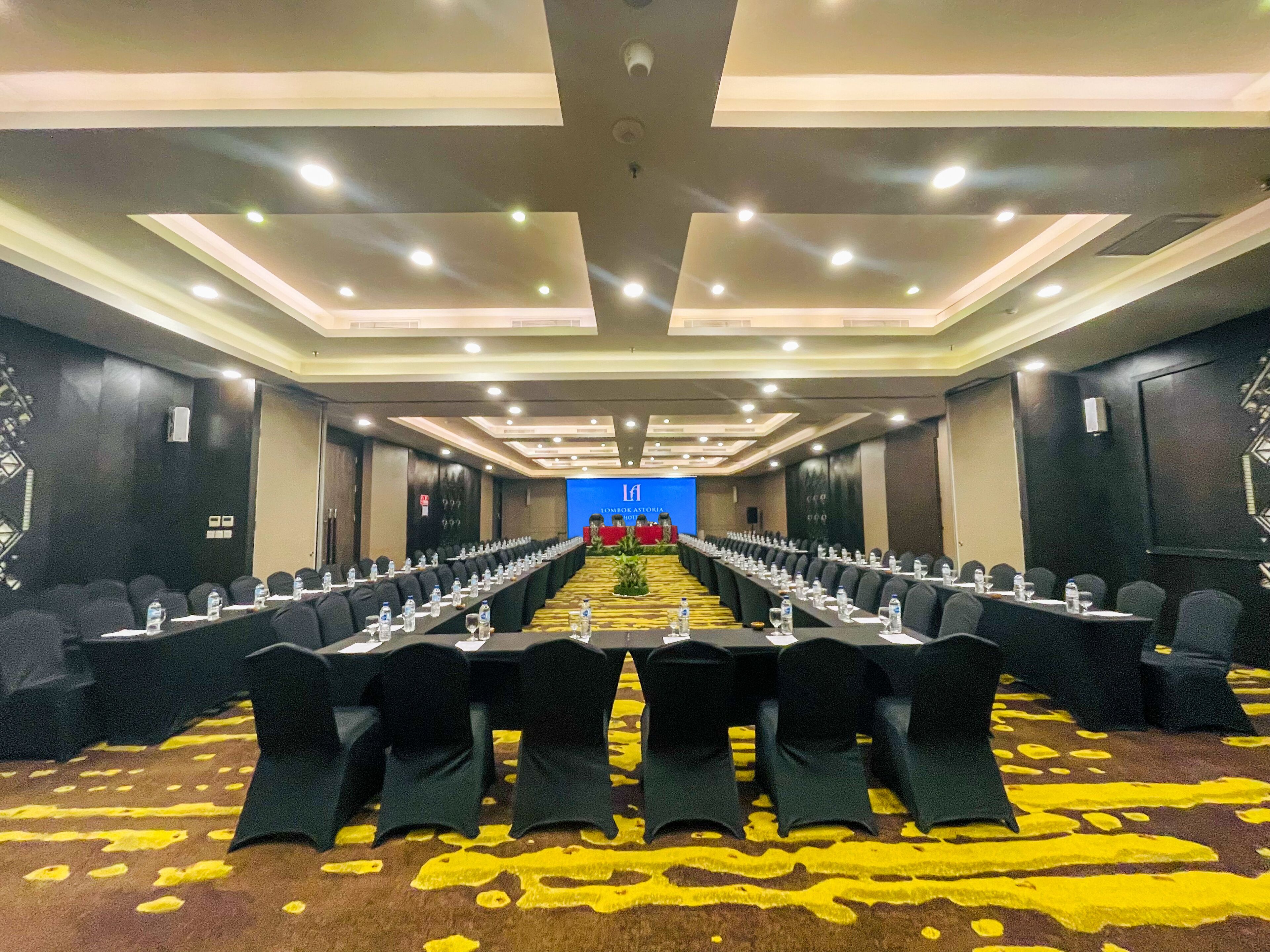 A conference room with rows of tables and chairs, a carpeted floor, and a blue screen at the front.