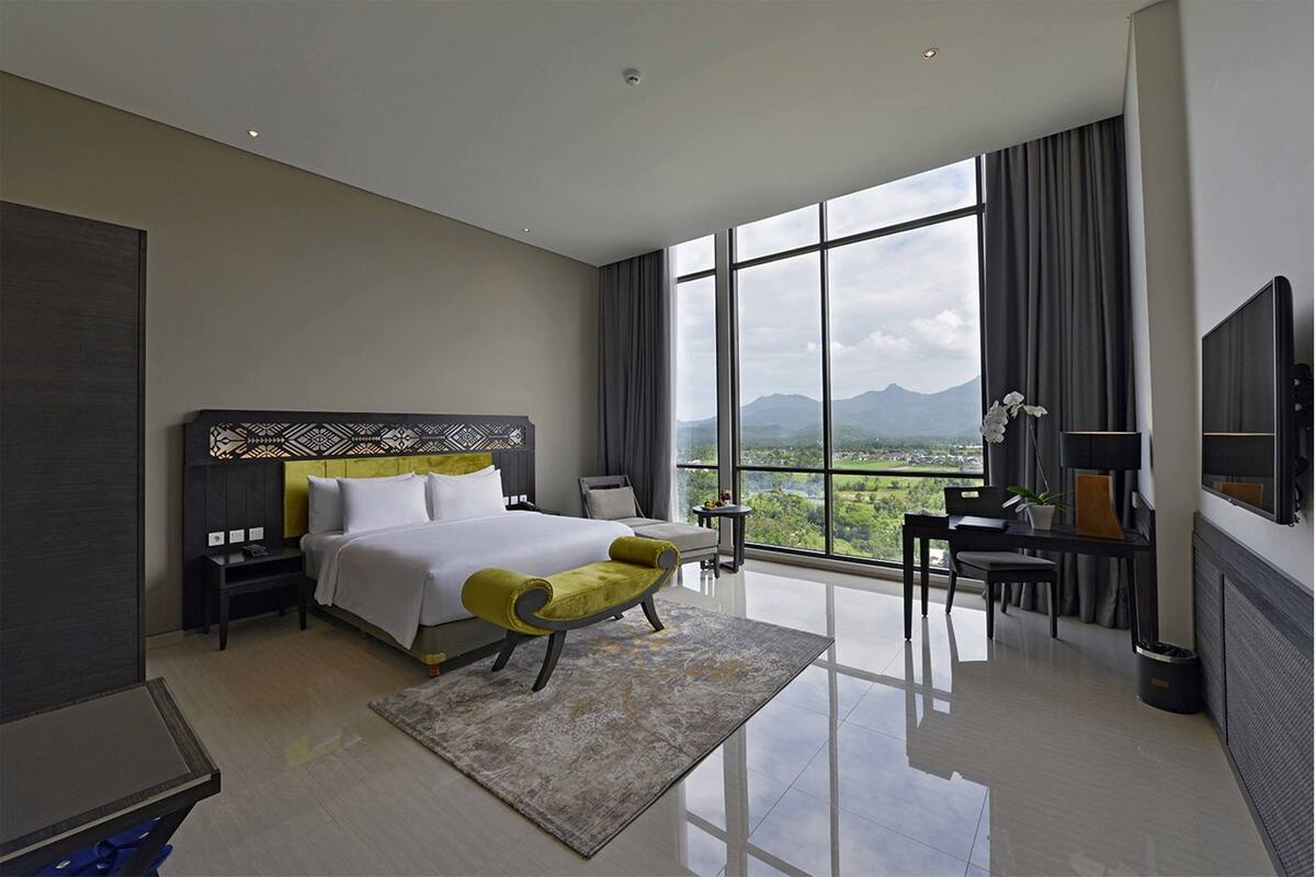 Ein modernes Hotelzimmer mit einem großen Bett, einer Sitzecke mit einem Sessel, einem Schreibtisch und Bergblick durch große Fenster.