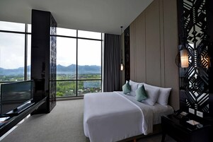 Habitación de hotel con una cama grande, televisor y vistas a montañas a través de amplios ventanales.