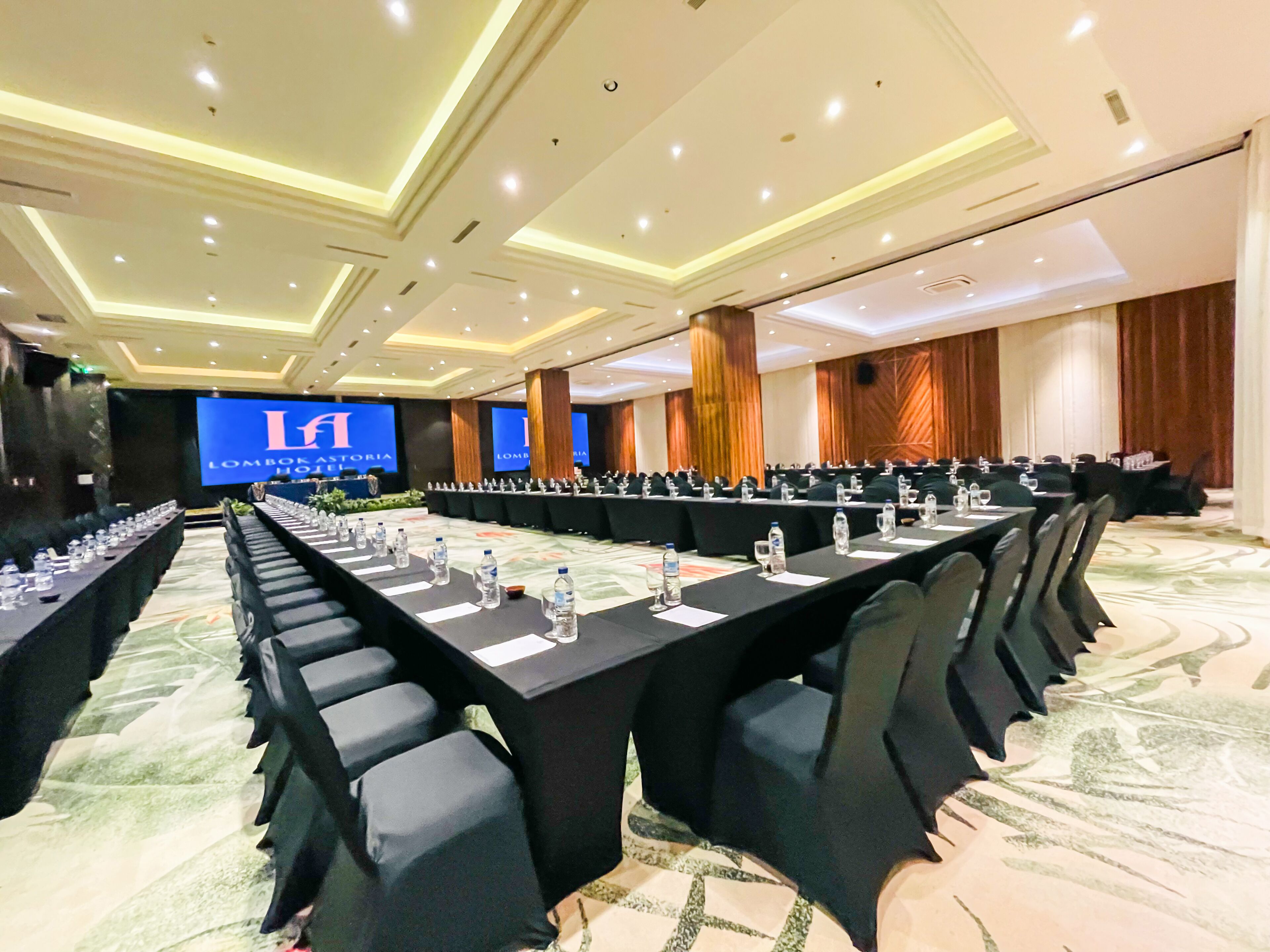A conference room with long tables, chairs, and a large screen displaying 'Lombok Astoria Hotel'.