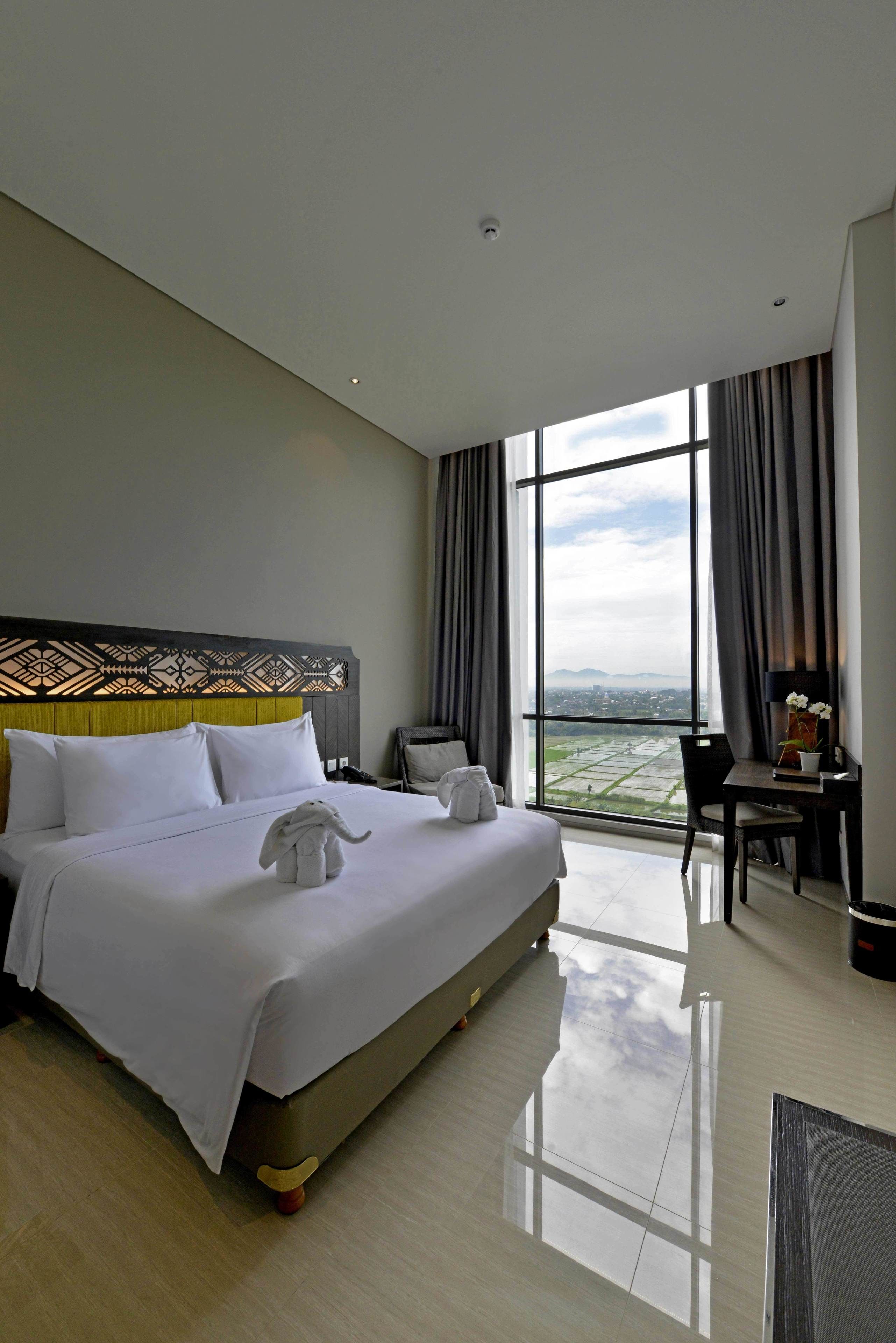 A hotel room with a large bed, a desk, and a view of the cityscape through a large window.