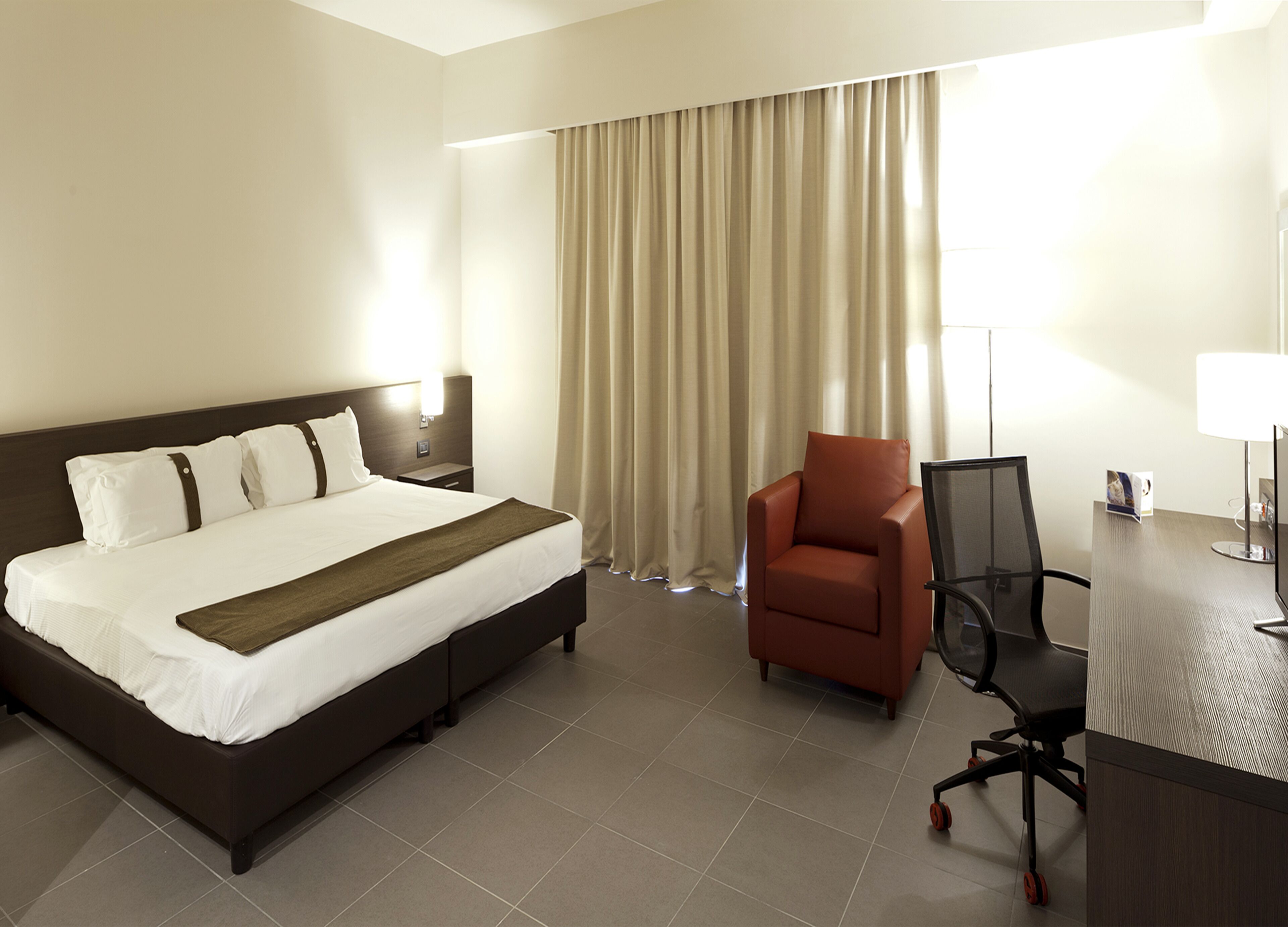 Una habitación de hotel con una cama grande, un escritorio con silla, una lámpara y una ventana con cortinas.