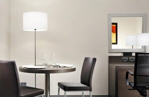 A hotel room with a round dining table, two chairs, a floor lamp, a mirror, and a minibar. - Inn Naples Airport (Gricignano d'Aversa)