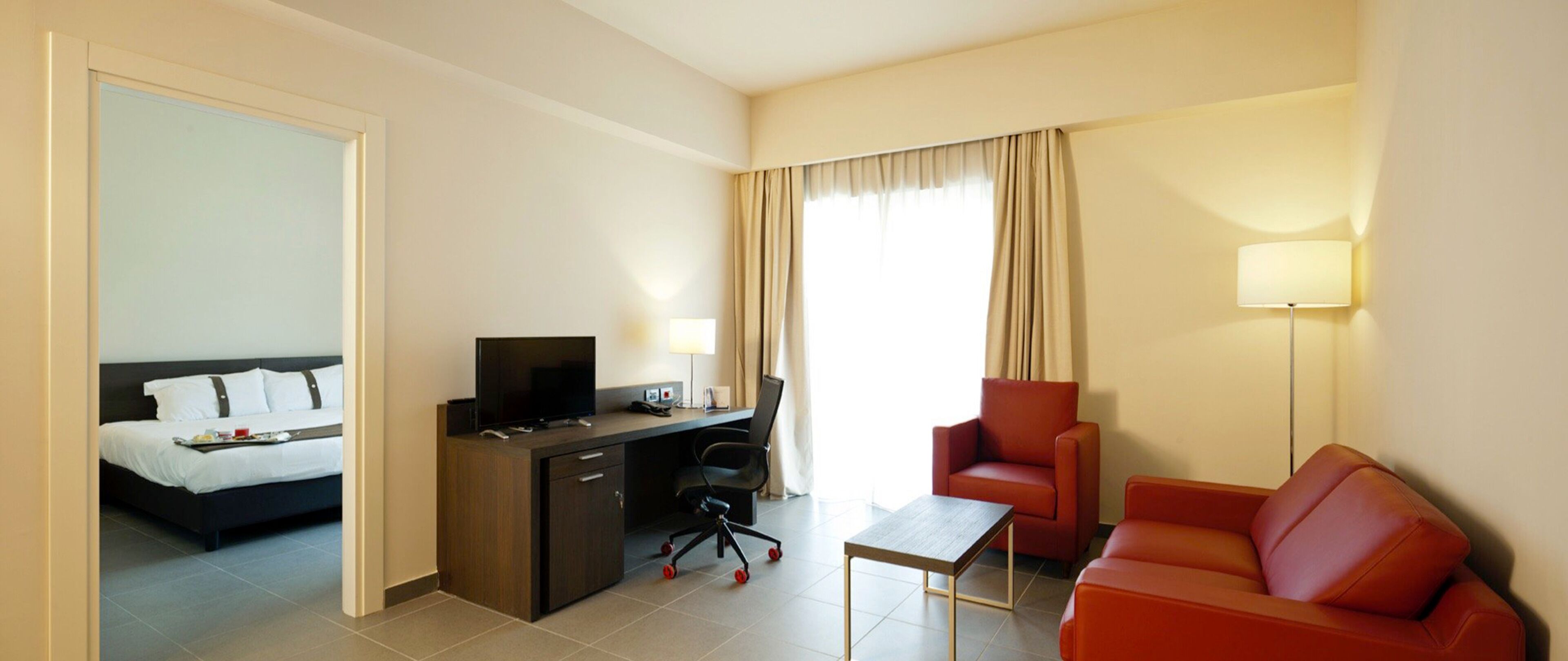 Quarto de hotel com cama, escrivaninha, computador, cadeira e uma pequena mesa.