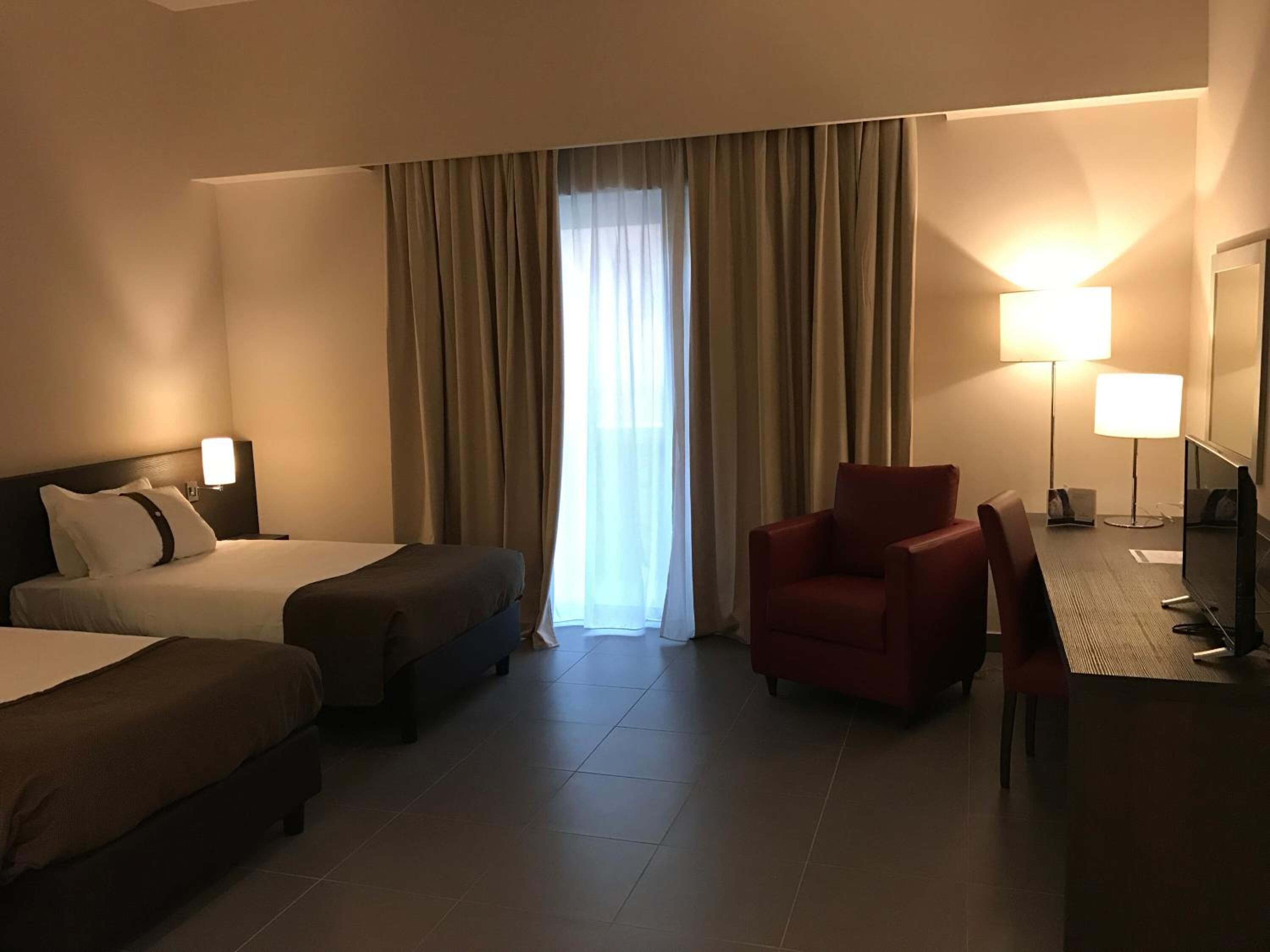 Habitació d'hotel amb dos llits, una taula de treball amb cadira, televisió i una finestra gran amb cortines.