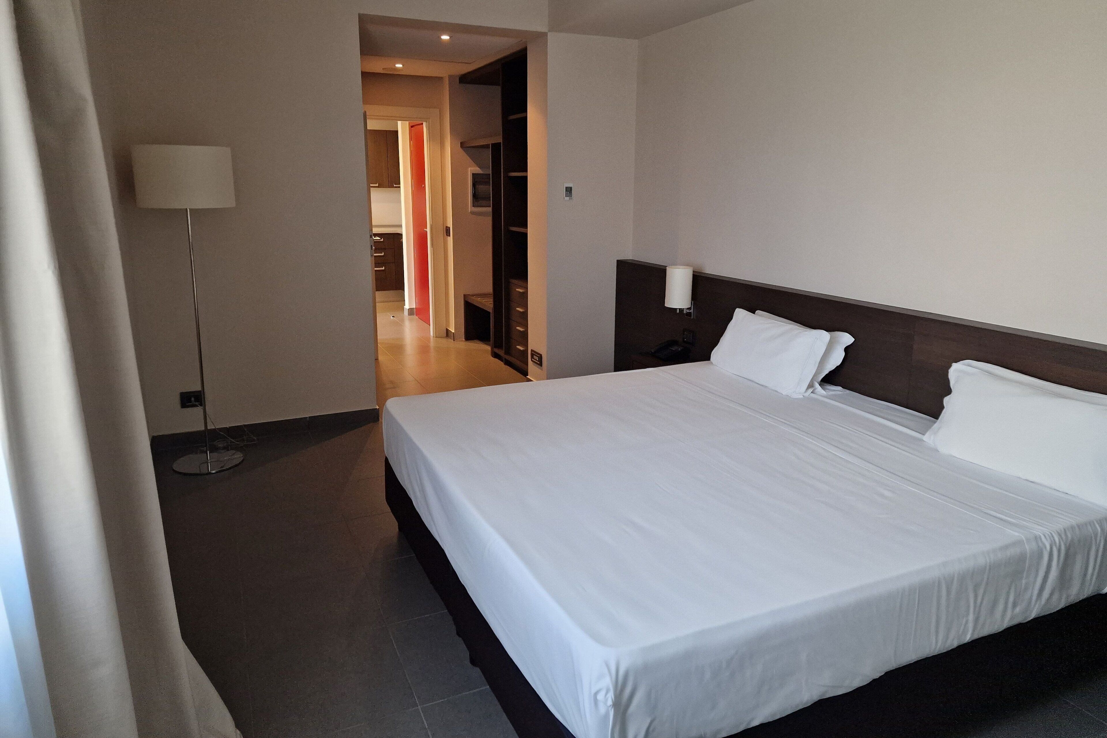 A hotel room with a large bed, a bedside table, a lamp, and a view of the hallway.