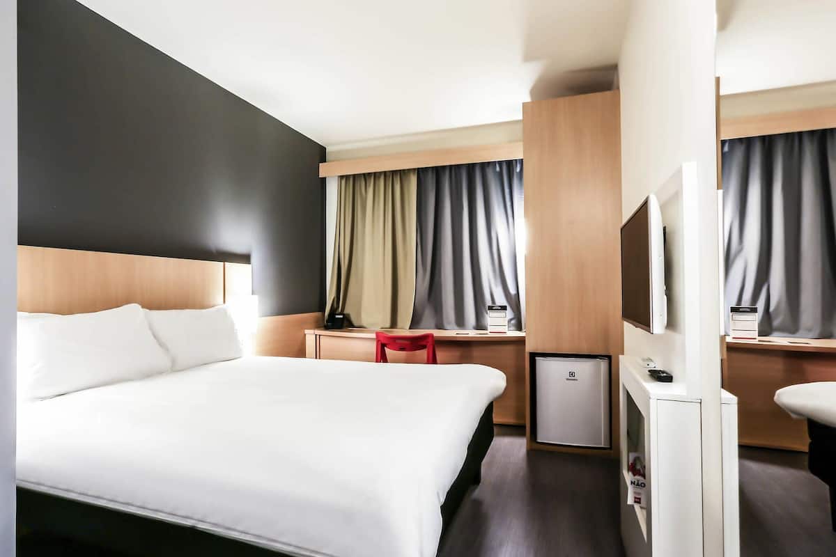 Quarto de hotel com cama, escrivaninha, cadeira, televisão e minibar.