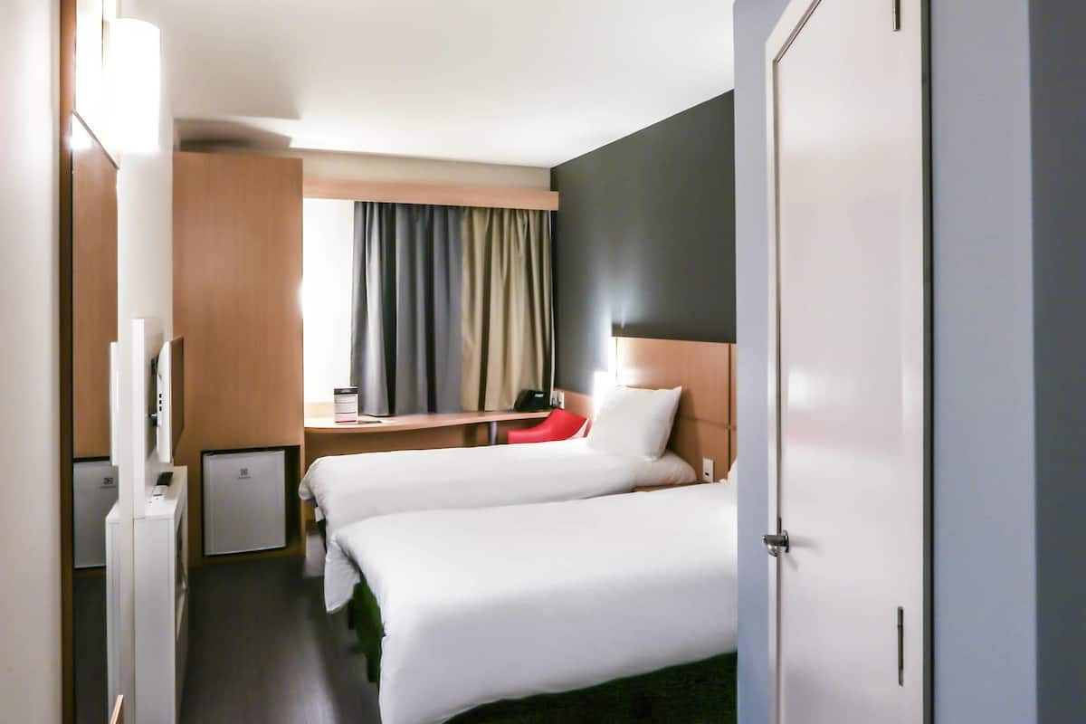 Quarto de hotel com duas camas, uma escrivaninha, uma televisão e uma janela com cortinas.