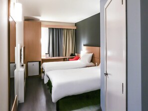 A hotel room with two beds, a desk, a TV, and a window with curtains.