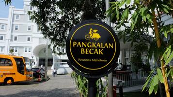 A sign for "Pangkalan Becak Pesona Maliboro" with a yellow bicycle icon, surrounded by greenery and a modern building in the background.