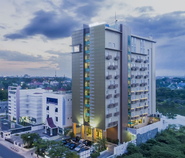 Un hôtel moderne, reconnaissable à son grand panneau « KHAS », doté d’un atrium central avec balcons, entouré d’une végétation luxuriante et comprenant un espace de stationnement.