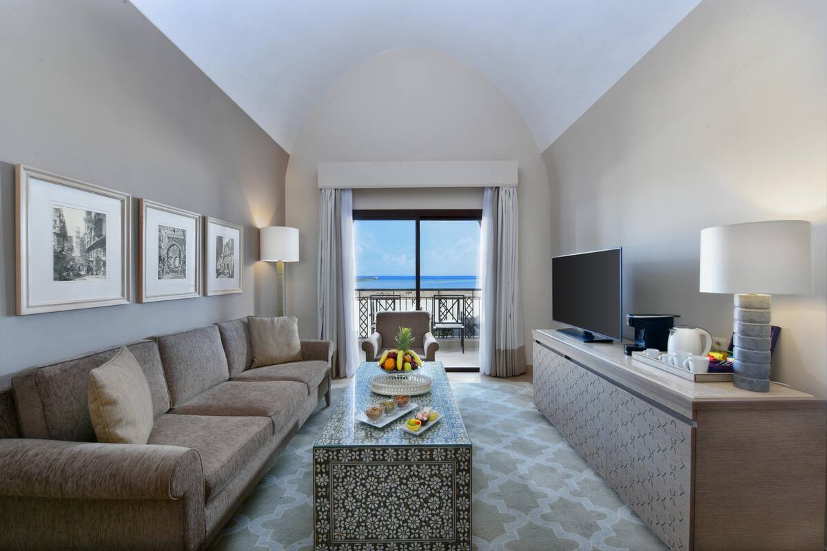 A hotel room with a balcony, a sofa, a coffee table with fruit and pastries, a TV, and framed pictures on the wall.