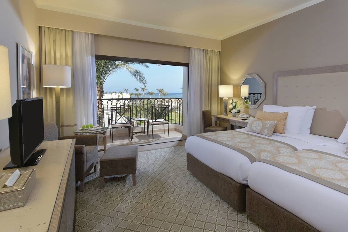 A hotel room with a bed, a desk with a computer, a chair, a small table with a plant, and a balcony with a view of the ocean.