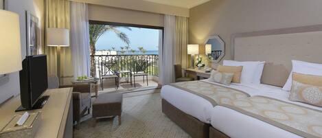 A hotel room with a bed, a desk with a computer, a chair, a small table with a plant, and a balcony with a view of the ocean.