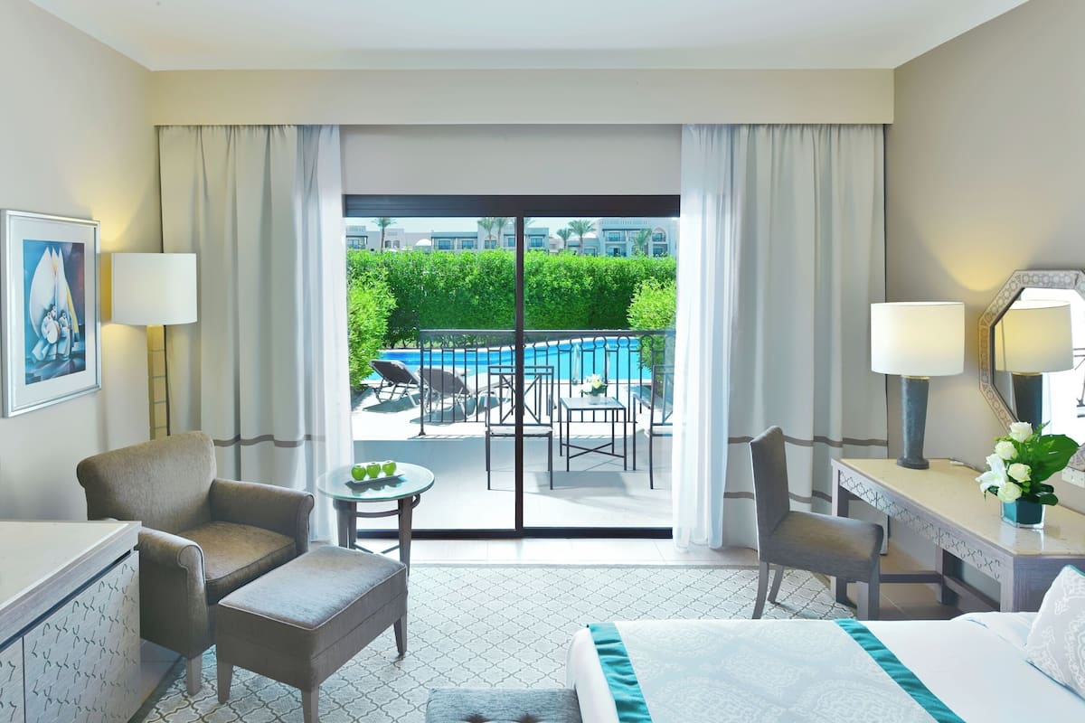 A hotel room with a bed, two armchairs, a desk, and a view of a pool and outdoor seating area.