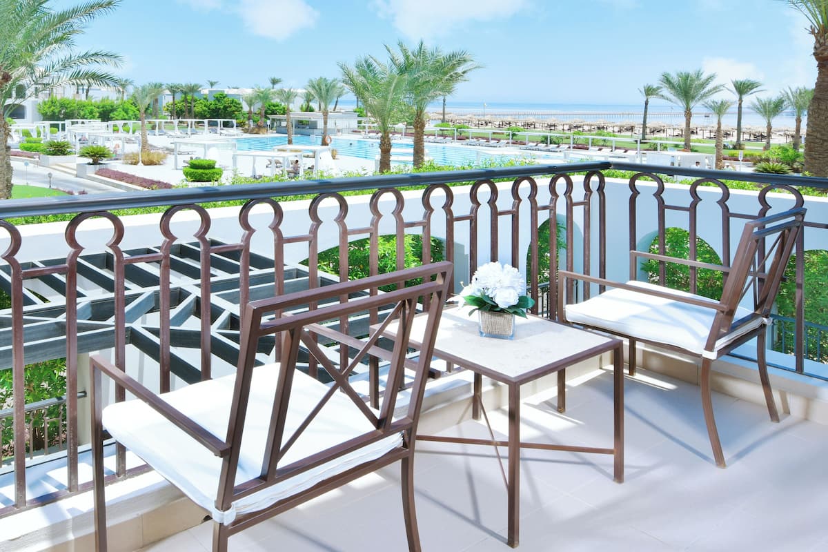 A balcony with a table and two chairs, overlooking a pool area with palm trees and a view of the sea.