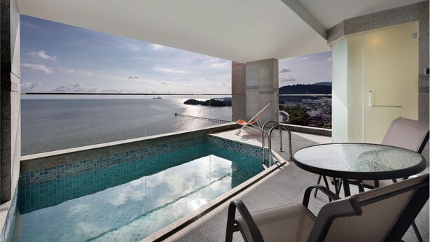 Un balcon moderne doté d’une piscine à débordement en verre, d’une table ronde et de chaises, offrant une vue sur une étendue d’eau et sur la terre ferme au loin.
