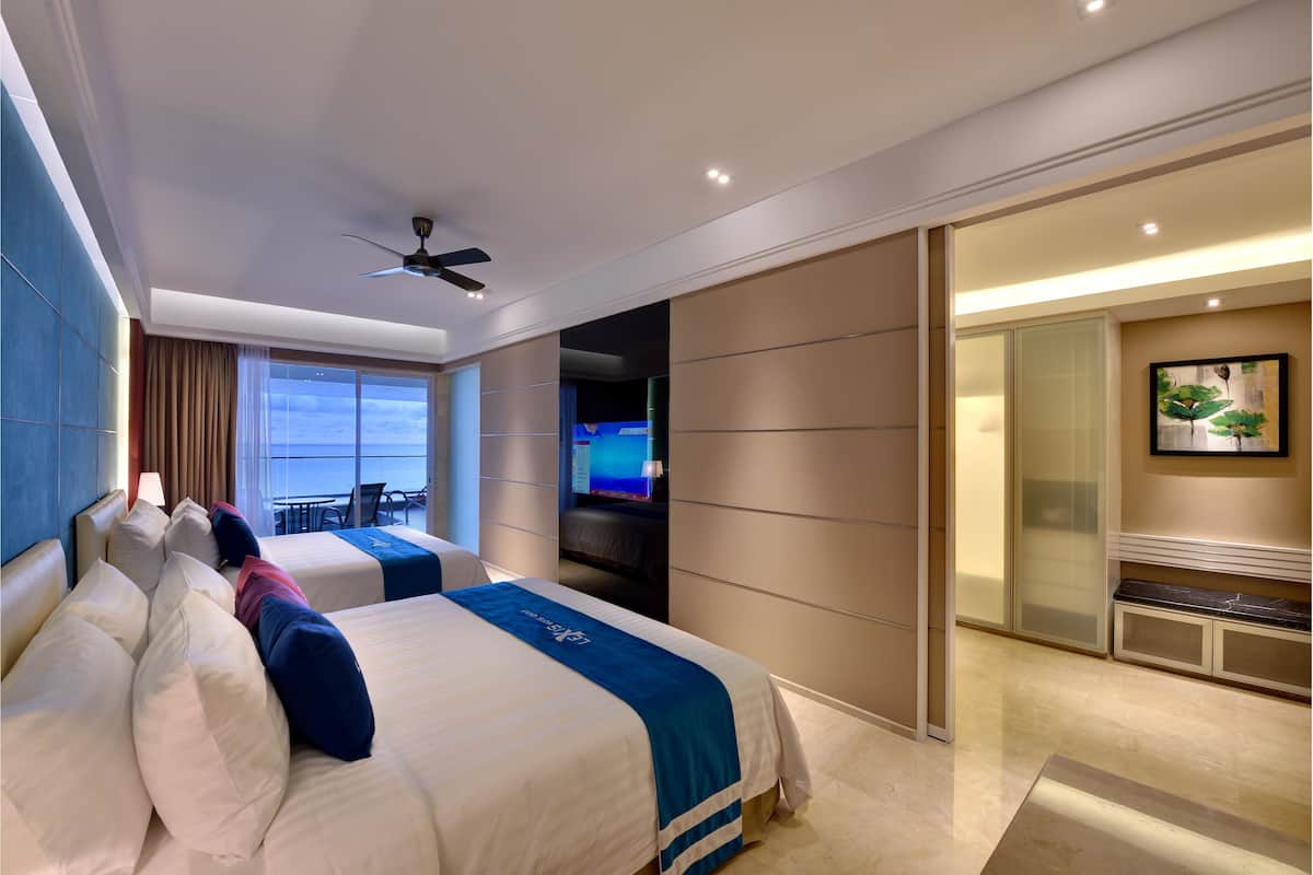 Habitación de hotel con dos camas, ventilador de techo, televisor y vista al océano.