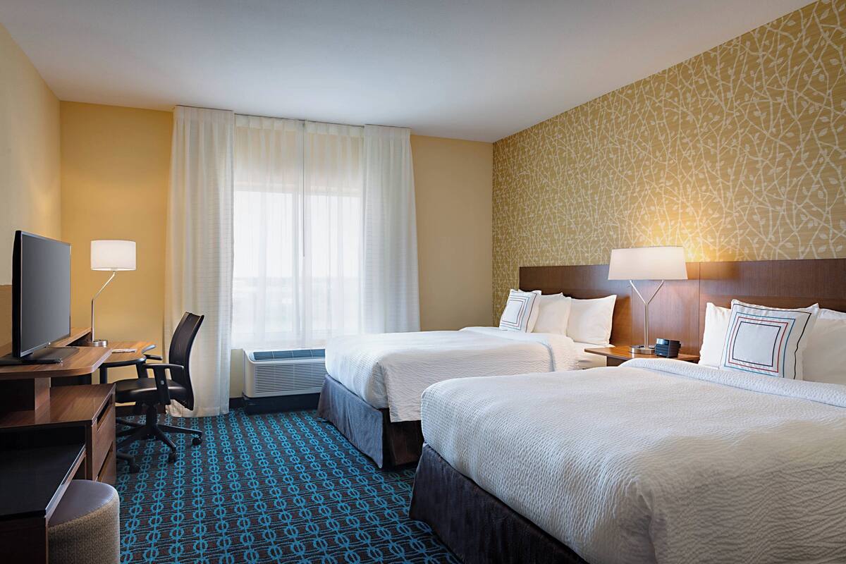 Ein Hotelzimmer mit zwei Betten, einem Schreibtisch, einem Fernseher, einem Sessel und einem fenster mit Vorhängen.