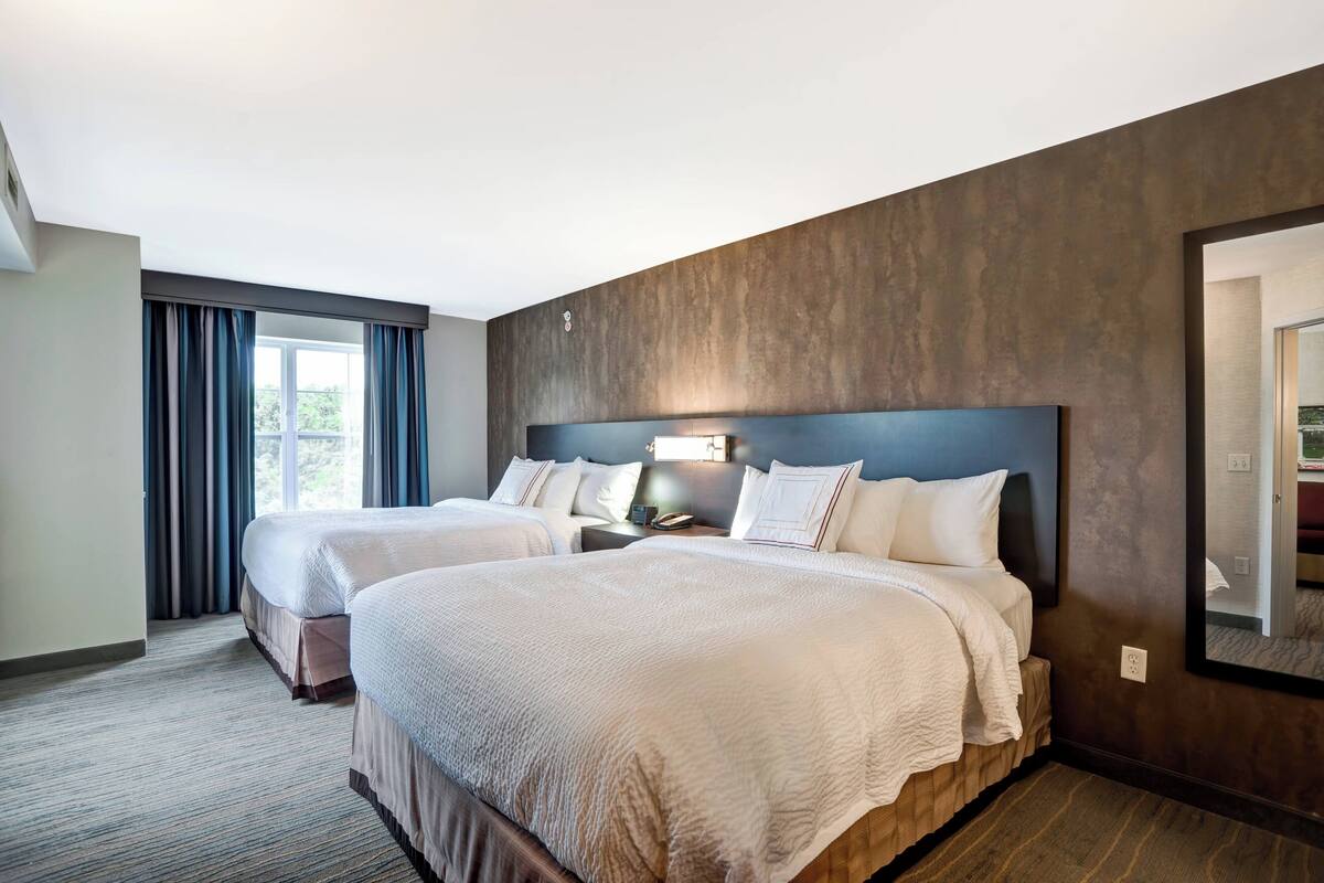 Une chambre d’hôtel avec deux lits, une grande fenêtre avec des rideaux, un téléviseur fixé au mur et un miroir.