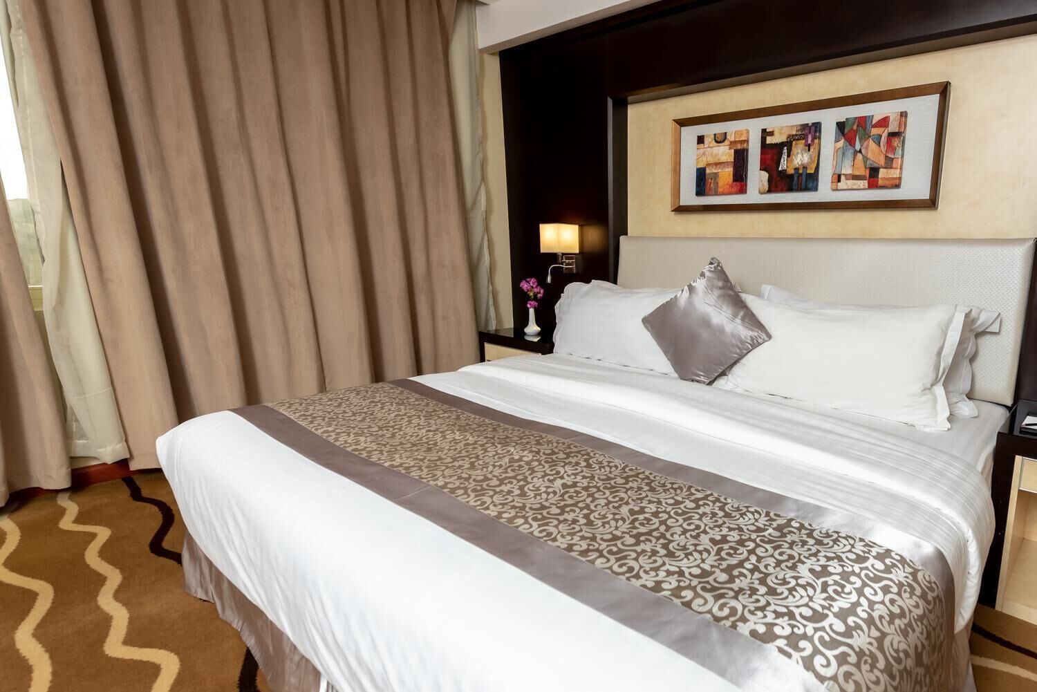 A hotel room with a large bed, beige curtains, a nightstand with a lamp, and abstract art on the wall.