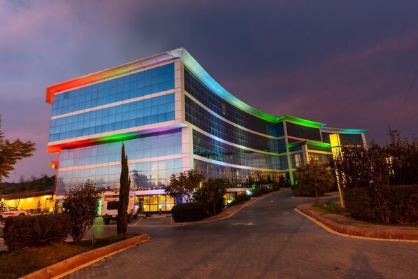 A modern hotel with a curved glass facade, a well-maintained driveway, and landscaped areas with shrubs and trees.