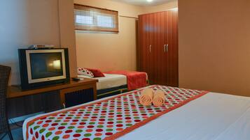 Quarto de hotel com uma cama, televisão sobre suporte de madeira, guarda-roupa e janela com persianas.