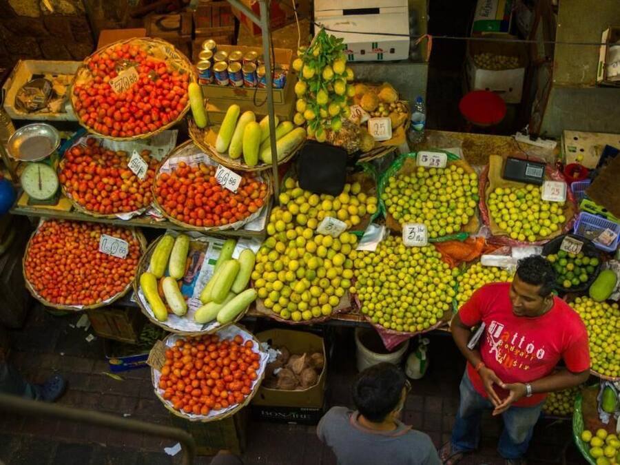 Un puesto de mercado con diversas frutas y verduras expuestas en cestas y cajas, con etiquetas de precios visibles.